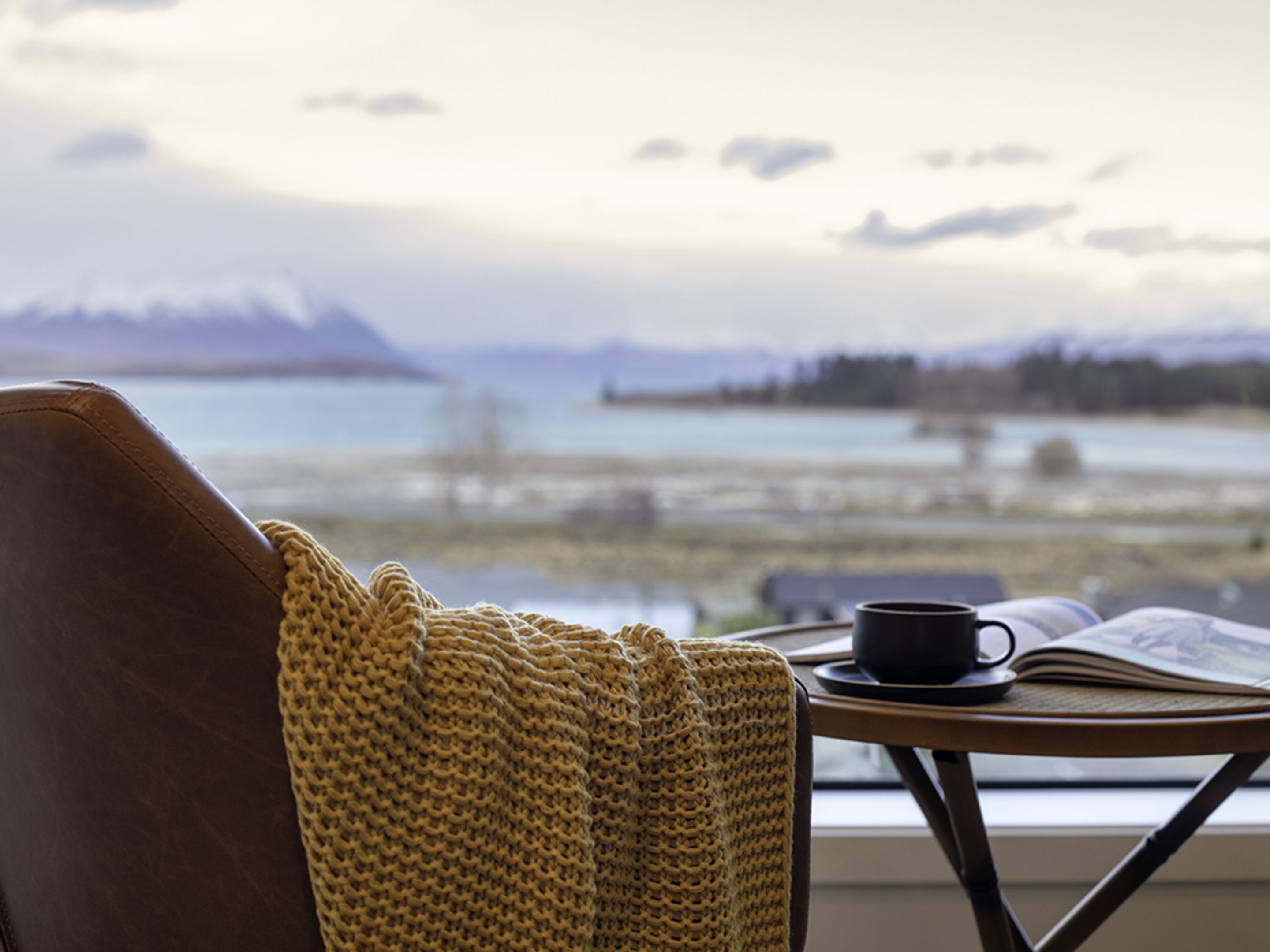 A view from a window with a mountain in the background, a table with a book and a cup, and a knitted blanket on a chair.