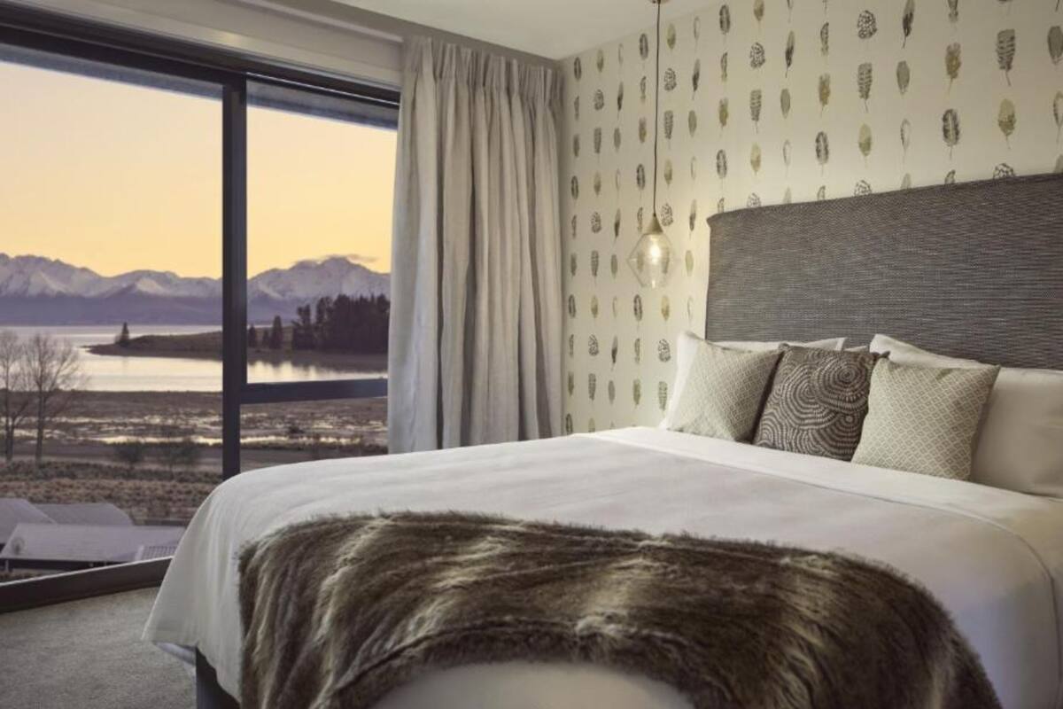 A modern hotel room with a large bed, a view of mountains and a lake, and a patterned wallpaper.