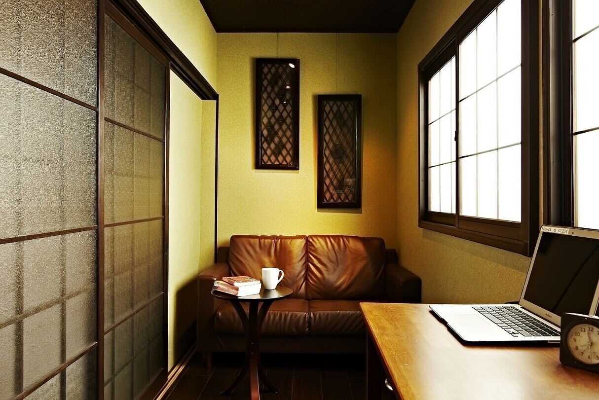 A cozy room with a brown leather sofa, a wooden table with a laptop and a clock, and a window with blinds.