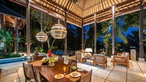 A covered outdoor dining area with a wooden table, chairs, and a pool in the background. - Villa Vajra (Ubud)