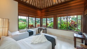 A hotel room with two beds, a ceiling fan, large windows with a view of greenery, and a small table with a lamp. - Villa Vajra (Ubud)
