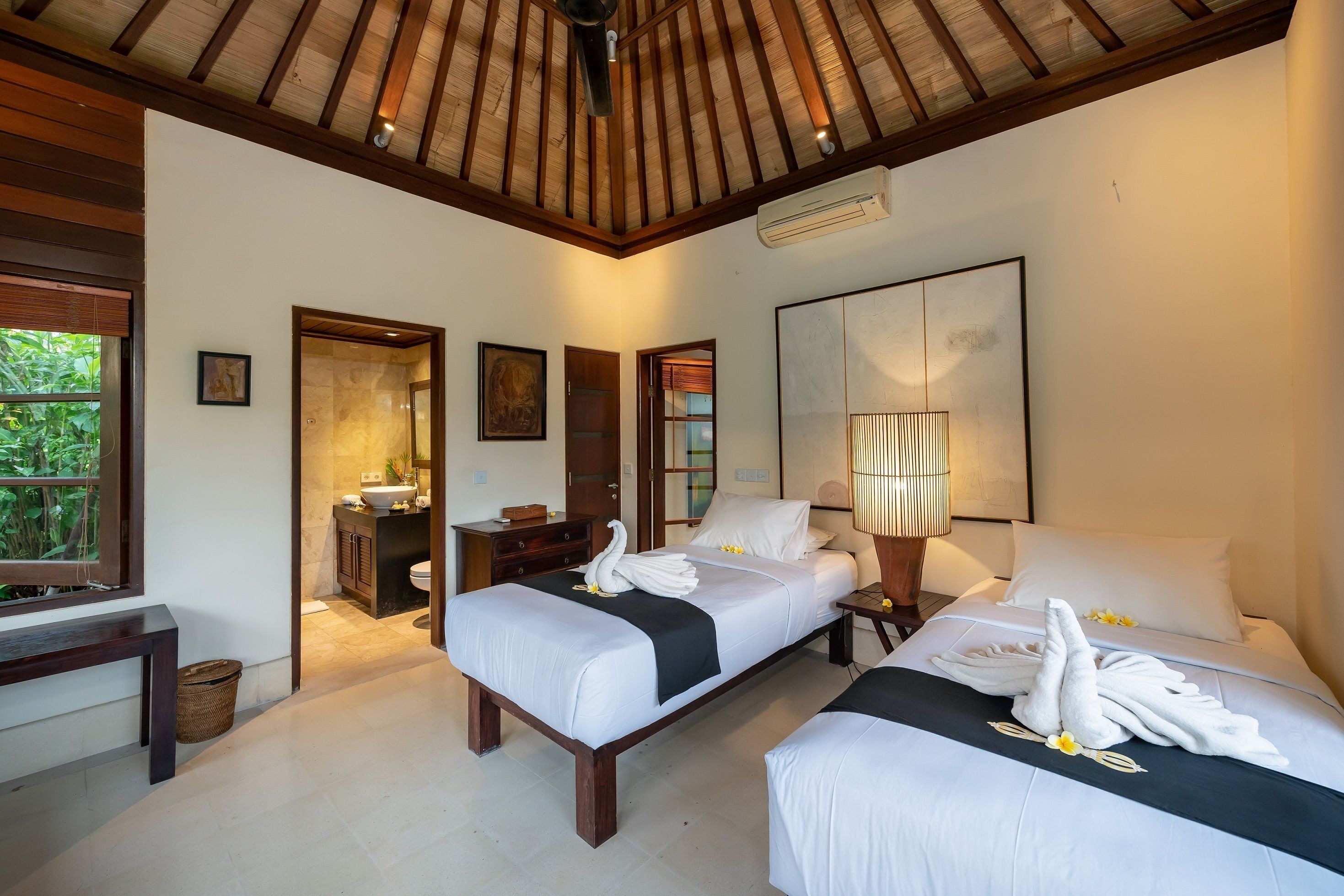 A hotel room with two beds, a wooden ceiling, a large window, a bathroom, and a painting on the wall.