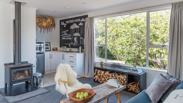 Un salon chaleureux avec une cheminée en pierre, un canapé, une table en bois sur laquelle se trouve un panier de pommes, et une vue sur l’extérieur grâce à de grandes fenêtres.