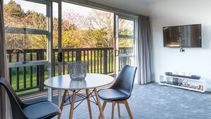 Un salon moderne avec une table ronde, deux chaises, un téléviseur et un balcon offrant une vue sur des arbres et une pelouse.