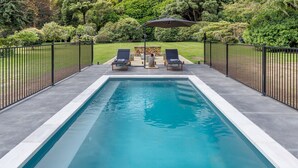 Une piscine rectangulaire, agrémentée de deux chaises longues et d’une table placée sous un auvent, le tout entouré d’une clôture en métal noir et d’une végétation luxuriante.