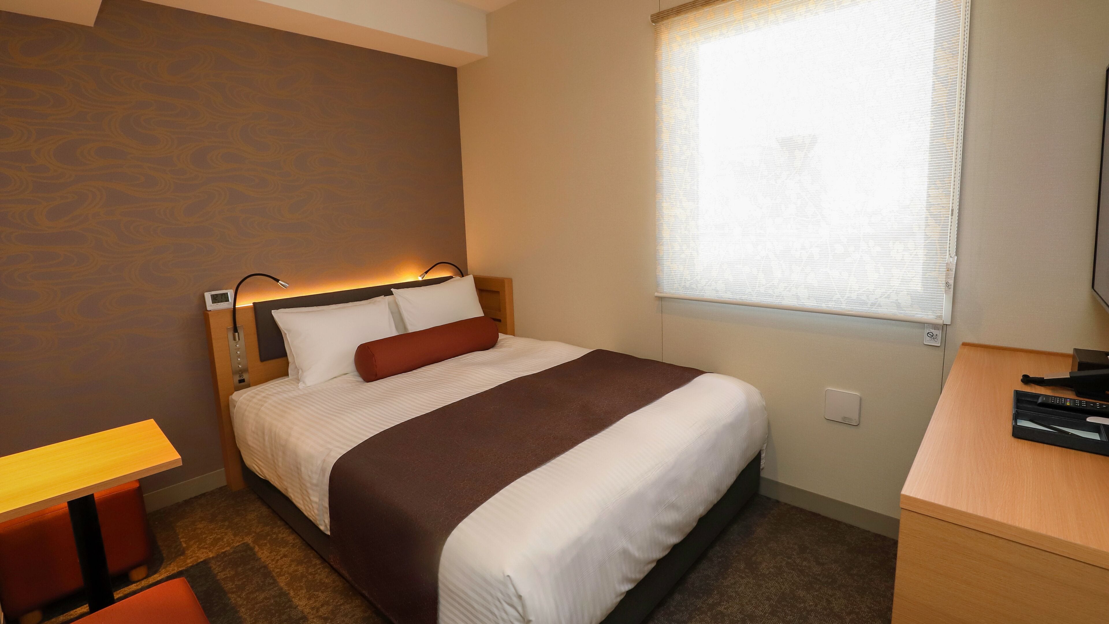 A hotel room with a bed, bedside table, lamp, and window with blinds.