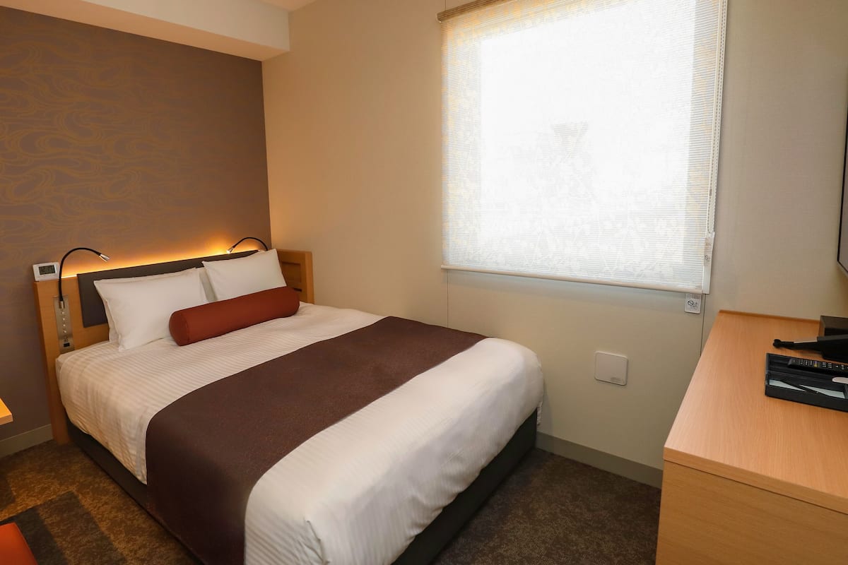 Una habitación de hotel con cama, mesita de noche, lámpara y ventana con persianas.
