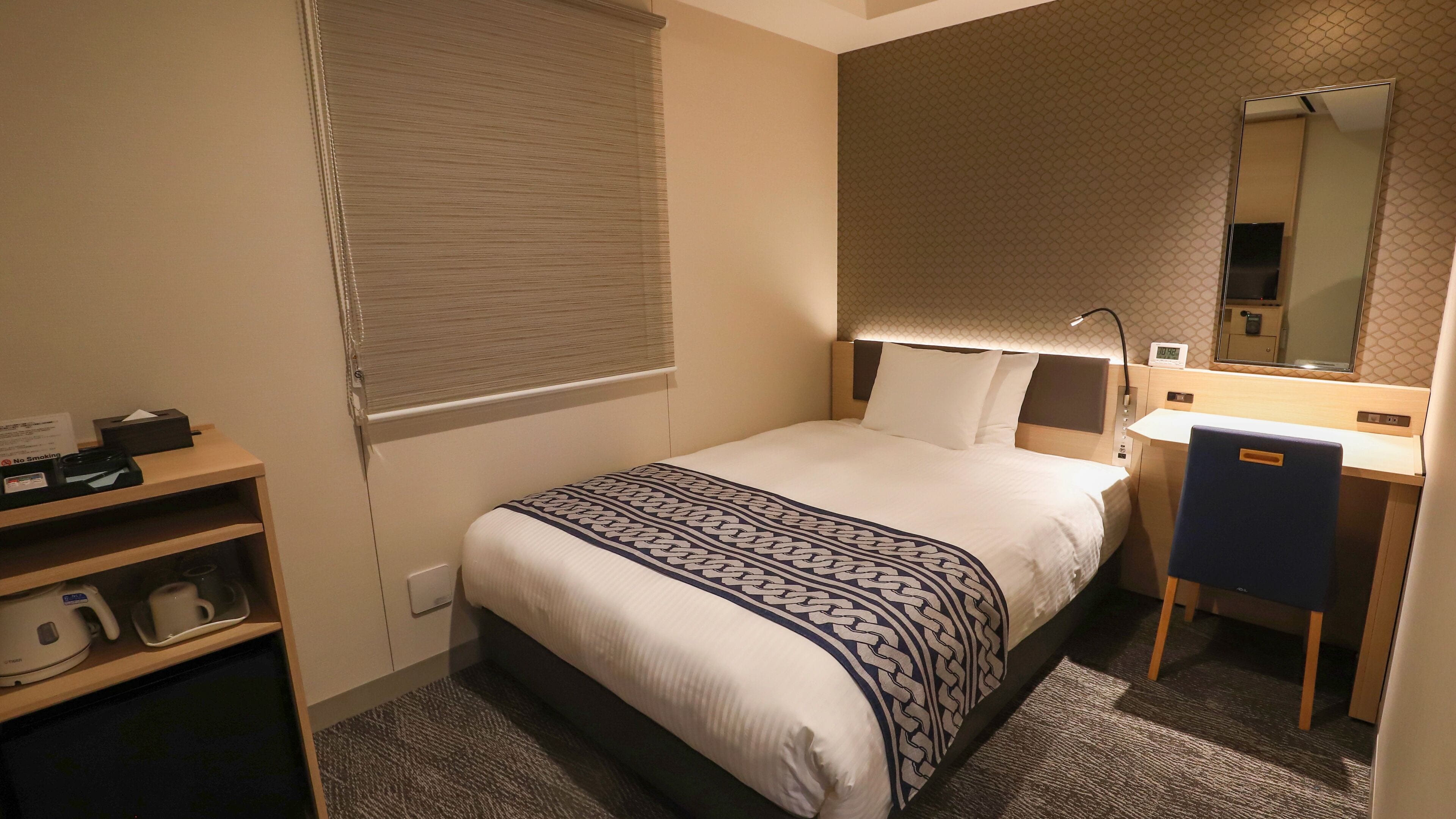 A hotel room with a bed, bedside table, lamp, mirror, and a small desk with a chair.
