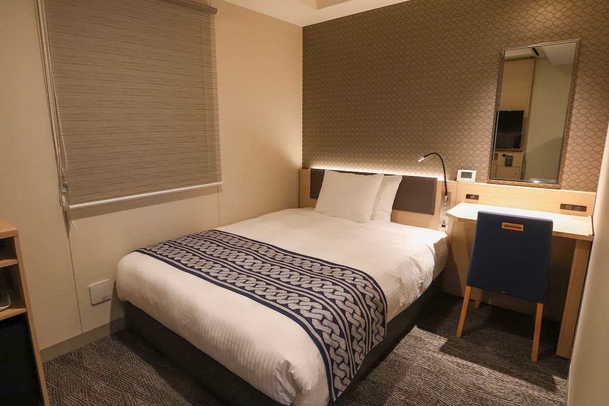 Una habitación de hotel con cama, mesita de noche, lámpara, espejo y un escritorio pequeño con una silla.