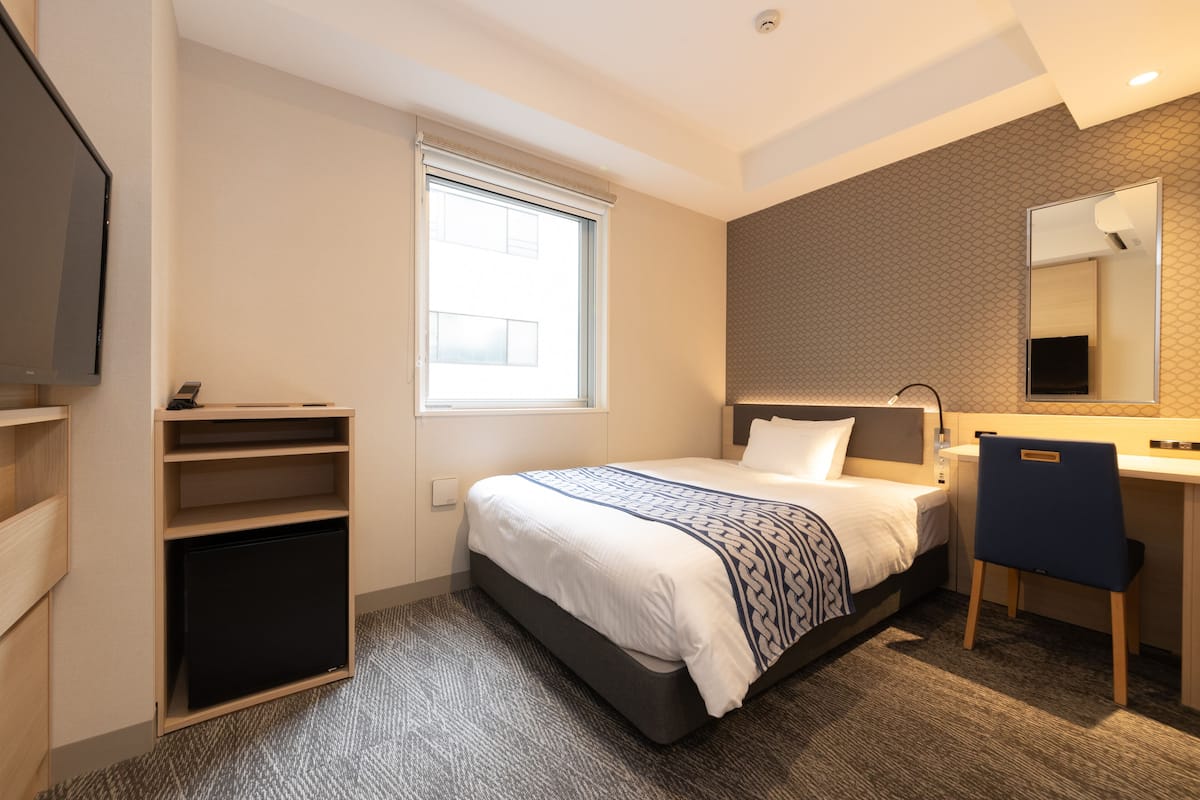 Una habitación de hotel con cama, escritorio, silla, televisor, armario y ventana.