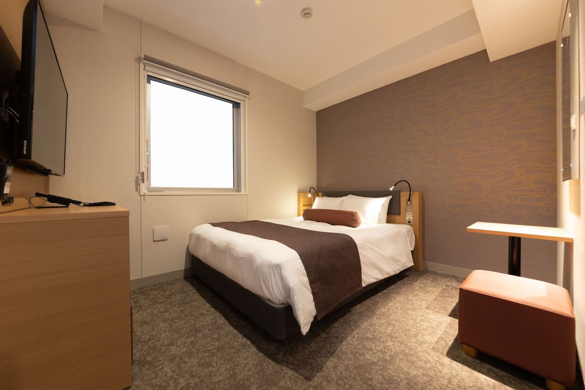 Una habitación de hotel moderna con una cama grande, un escritorio, un televisor y una ventana que proporciona luz natural.