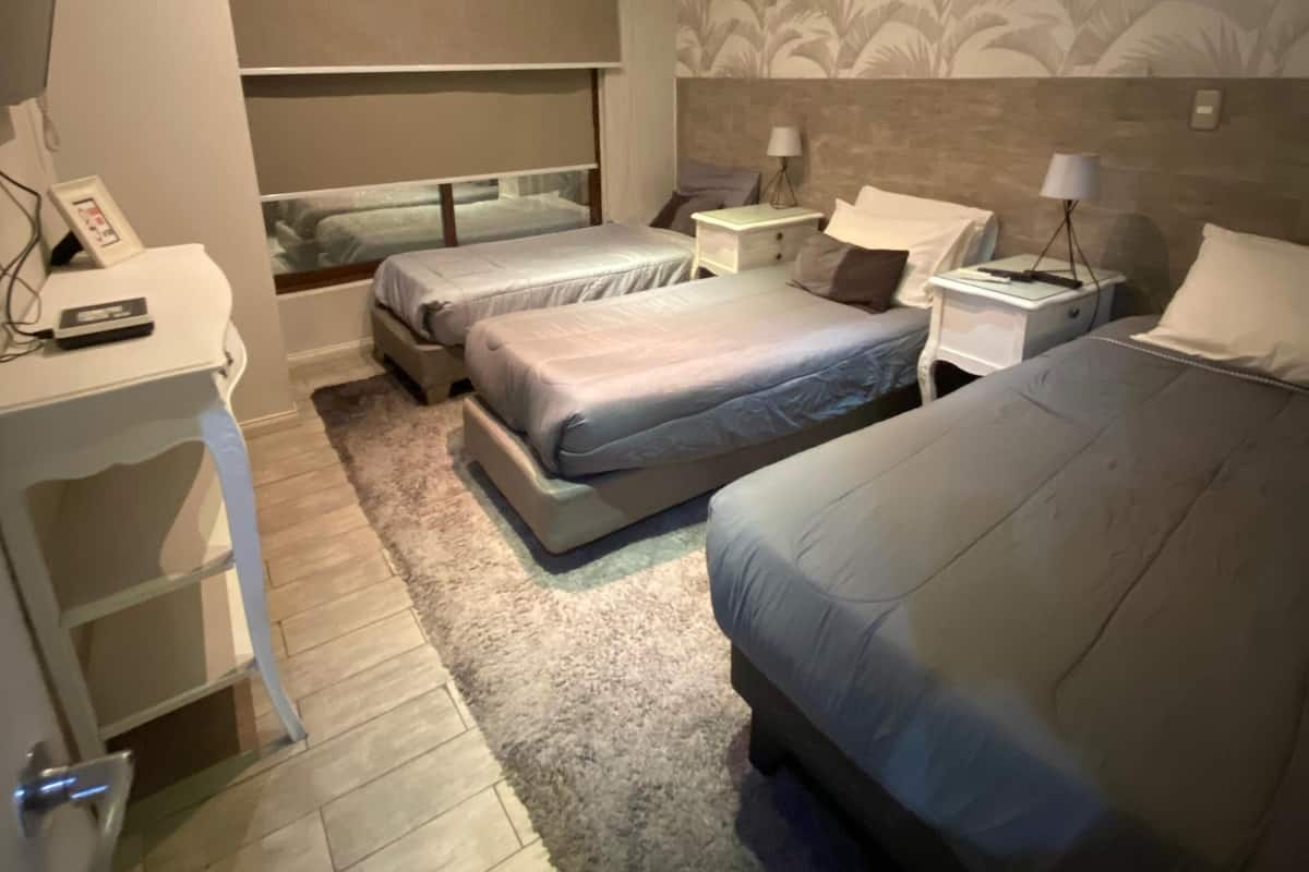 A hotel room with two beds, a desk, a chair, a bedside table with a lamp, and a wall-mounted air conditioner.