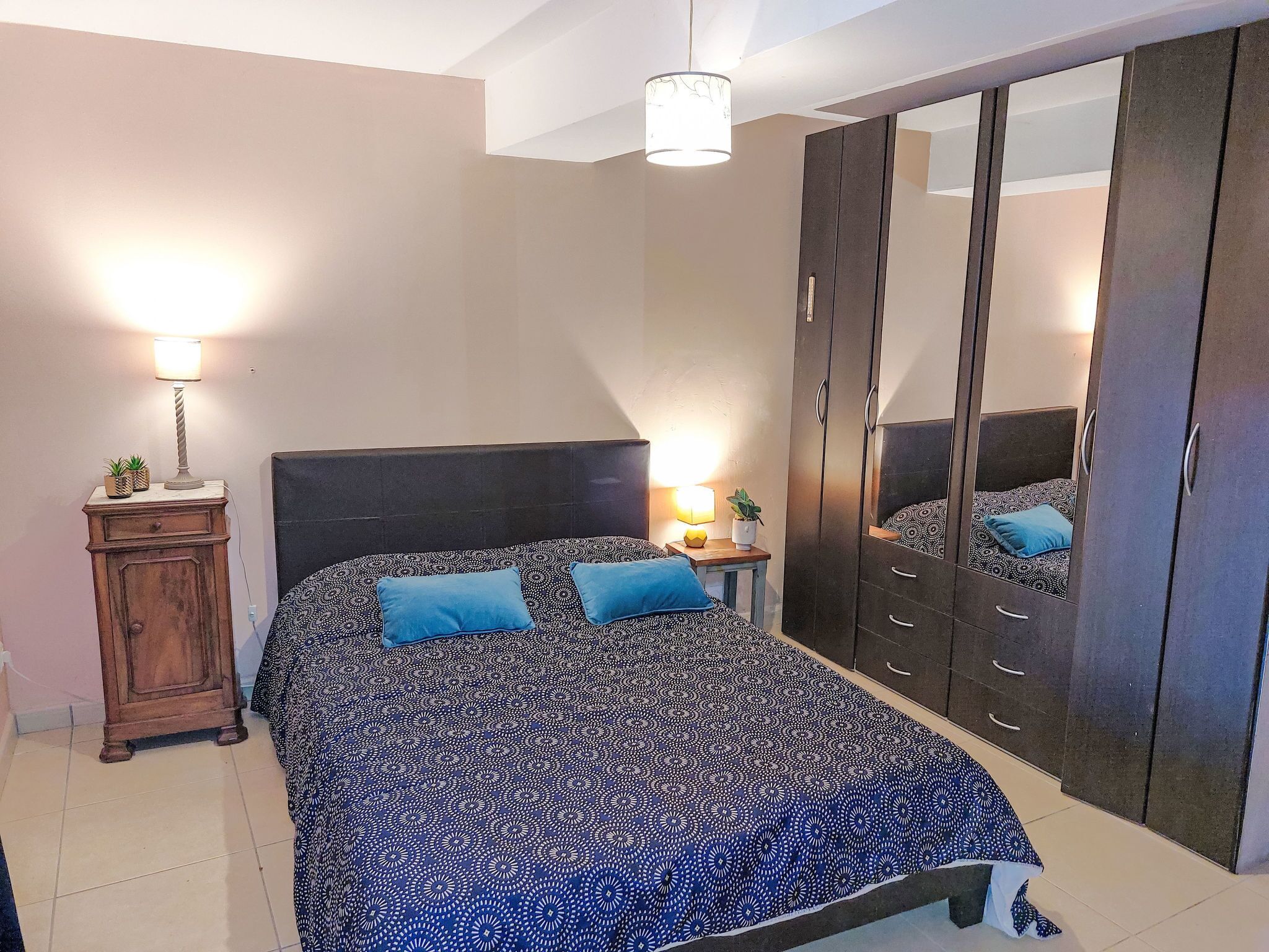 A bedroom with a patterned bedspread, a wooden wardrobe, a bedside table with a lamp, and a mirror on the wardrobe.
