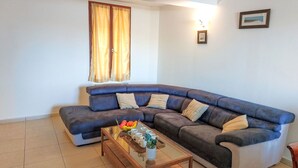 A living room with a blue sectional sofa, a glass coffee table with fruit and cups, and a window with yellow curtains. - La Maison de l'Etang by Interhome (Gruissan)