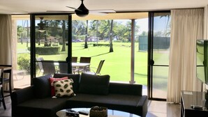 Una sala de estar con un sofá de color oscuro, una mesa de centro y un mueble para el televisor. Hay un amplio ventanal corredero que da a un área exterior verde con césped y árboles.