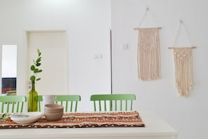Un comedor con una mesa blanca, sillas verdes, un mantel individual estampado, una botella verde y dos tapices de macramé.