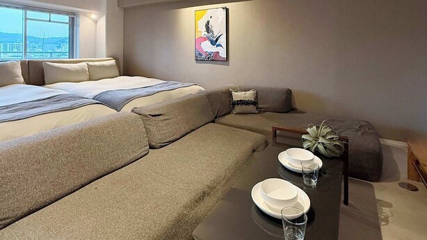 A modern living room with a sofa, a dining table with plates and glasses, a bed with pillows, and a window with a view. - ORIGAMINN 601 by b hotel - 5 mins PeacePark (Hiroshima City)