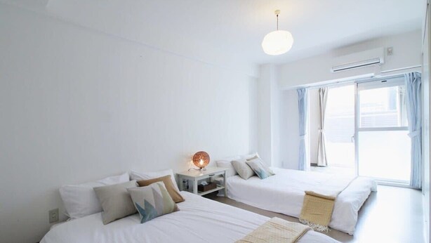 A hotel room with two beds, a bedside table with a lamp, a window with curtains, and an air conditioning unit.