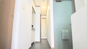 Un couloir étroit avec une porte en bois sur la gauche, un mur blanc avec un interrupteur, et une porte bleu clair au fond.