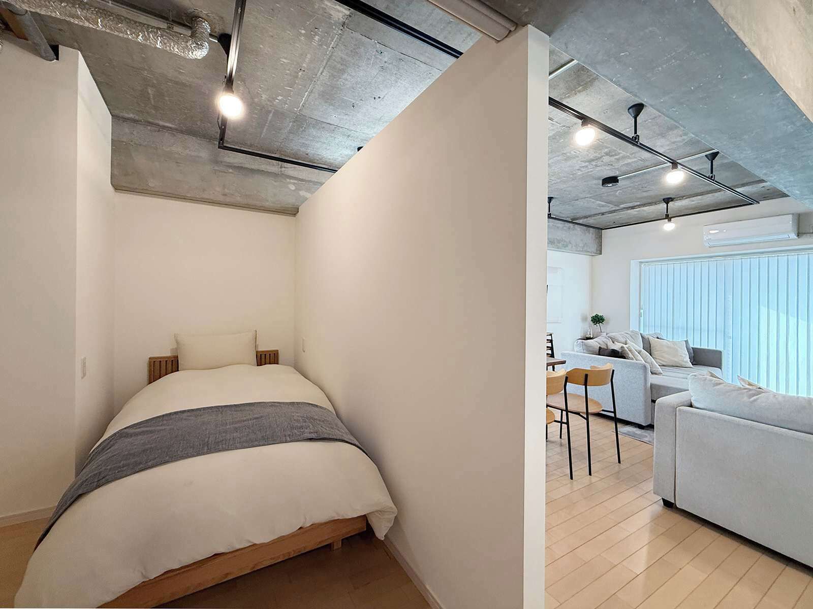 A modern bedroom with a bed, a small table, and a sofa in a bright living area with large windows.