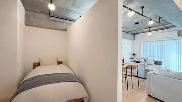 A modern bedroom with a bed, a small table, and a sofa in a bright living area with large windows.