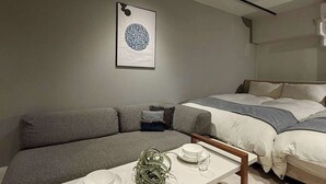 A modern bedroom with a gray sofa, a bed with white bedding, a bedside table with a glass and a bowl, and a framed artwork on the wall. - ORIGAMINN 502 by b hotel - 5 mins PeacePark (Hiroshima City)