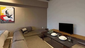 A modern living room with a sectional sofa, a flat-screen TV, a dining table with plates and glasses, and a decorative wall piece. - ORIGAMINN 501 by b hotel -  5 mins PeacePark (Hiroshima City)