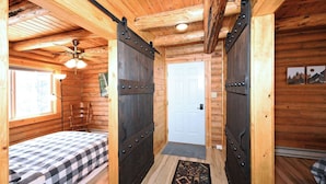 Habitación de cabaña de troncos con una cama, un ventilador de techo y una puerta con cerrojo de metal.