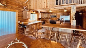 Interior rústico de una cabaña de madera con una gran mesa de comedor de madera, taburetes de bar, una zona de cocina y un techo con vigas de madera y cornamentas.