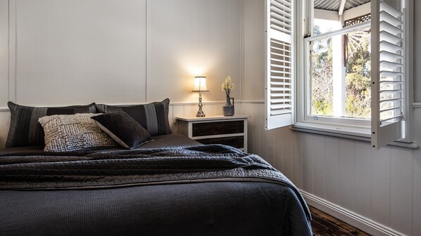A bedroom with a bed, bedside table, lamp, and window with shutters. - Table Hill Cottage is your quintessential Daylesford miner’s cottage (Daylesford)