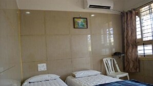 A room with two beds, a white chair, a window with curtains, and a wall-mounted air conditioner. - Mourya Deluxe AC Lodge (Chitradurga)