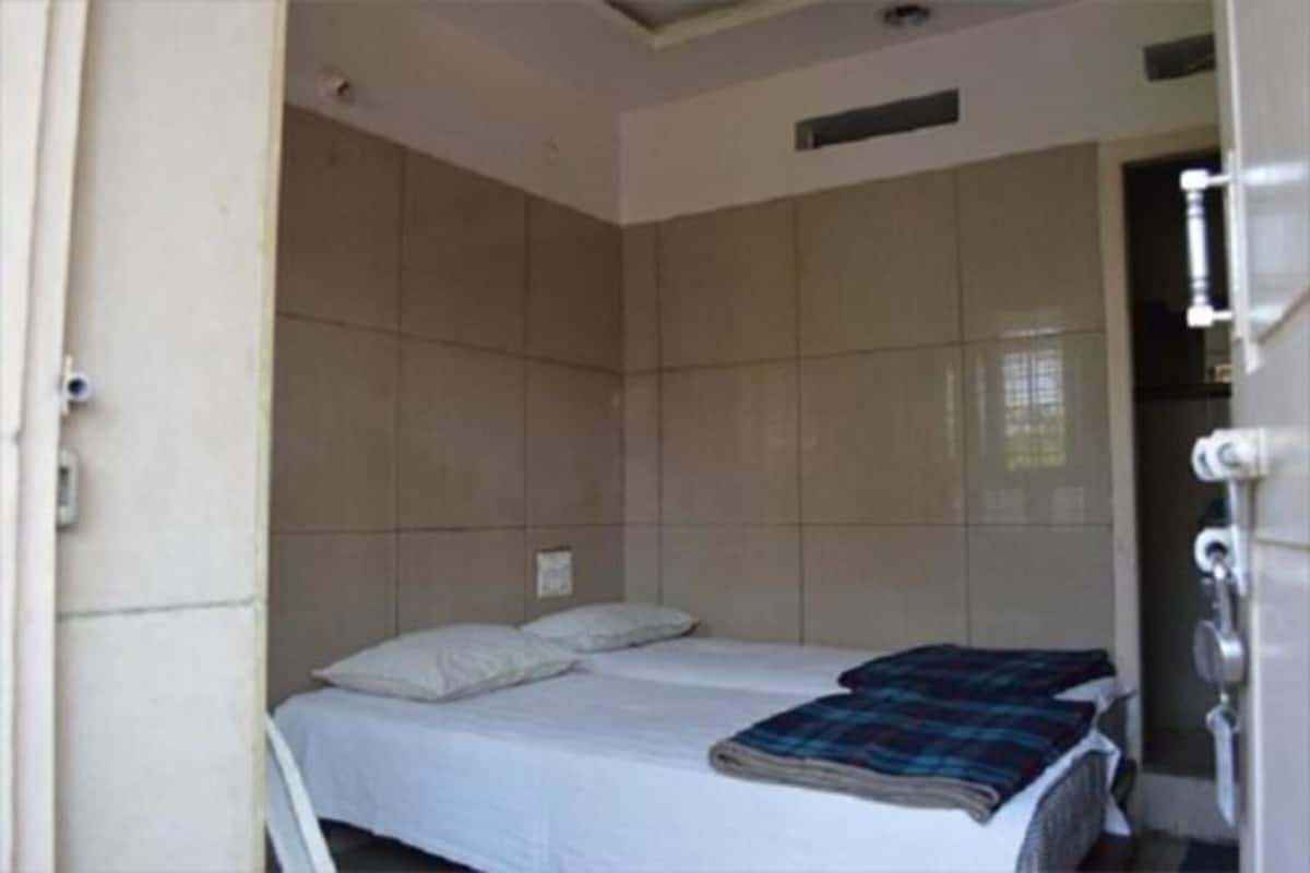 Une petite chambre avec un lit simple, un mur recouvert de carreaux unis, un lit avec du linge de lit blanc et une couverture à carreaux, et une porte de salle de bain visible.