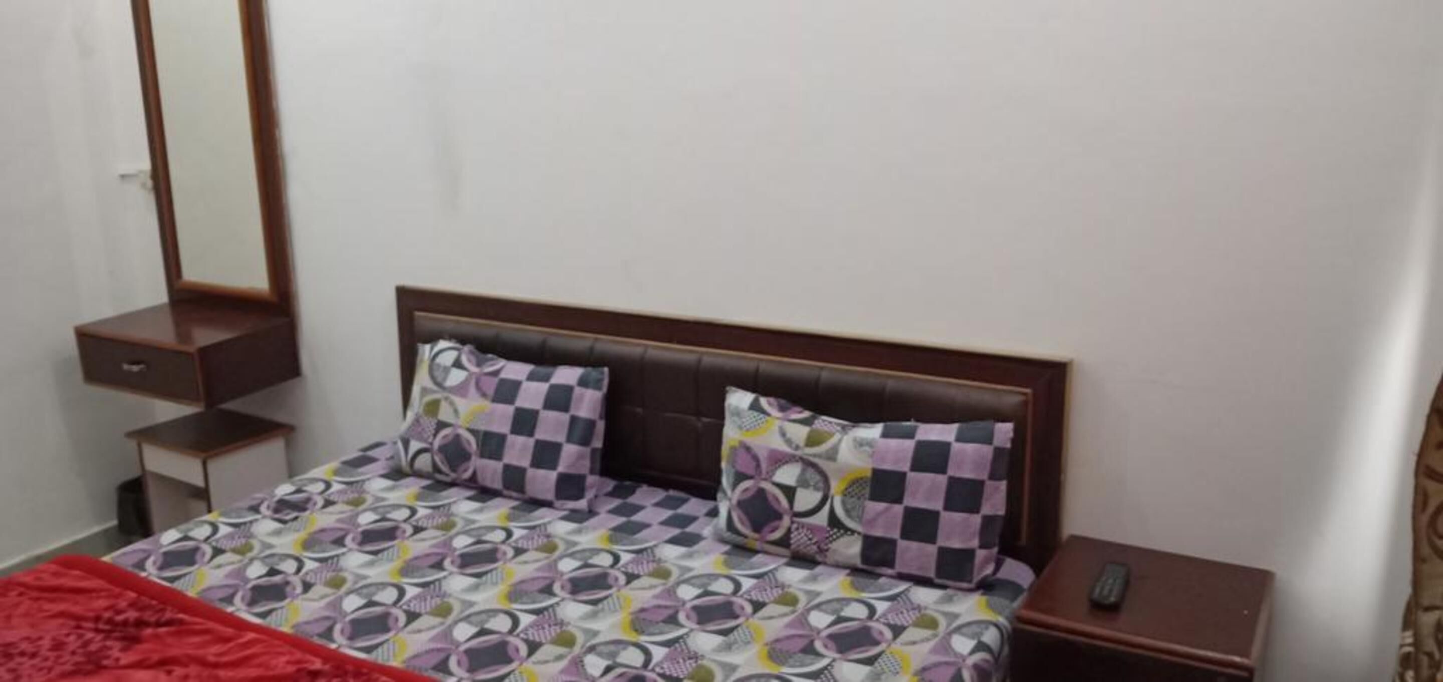 A bedroom with a bed, two pillows, a nightstand, a mirror, and a red blanket.