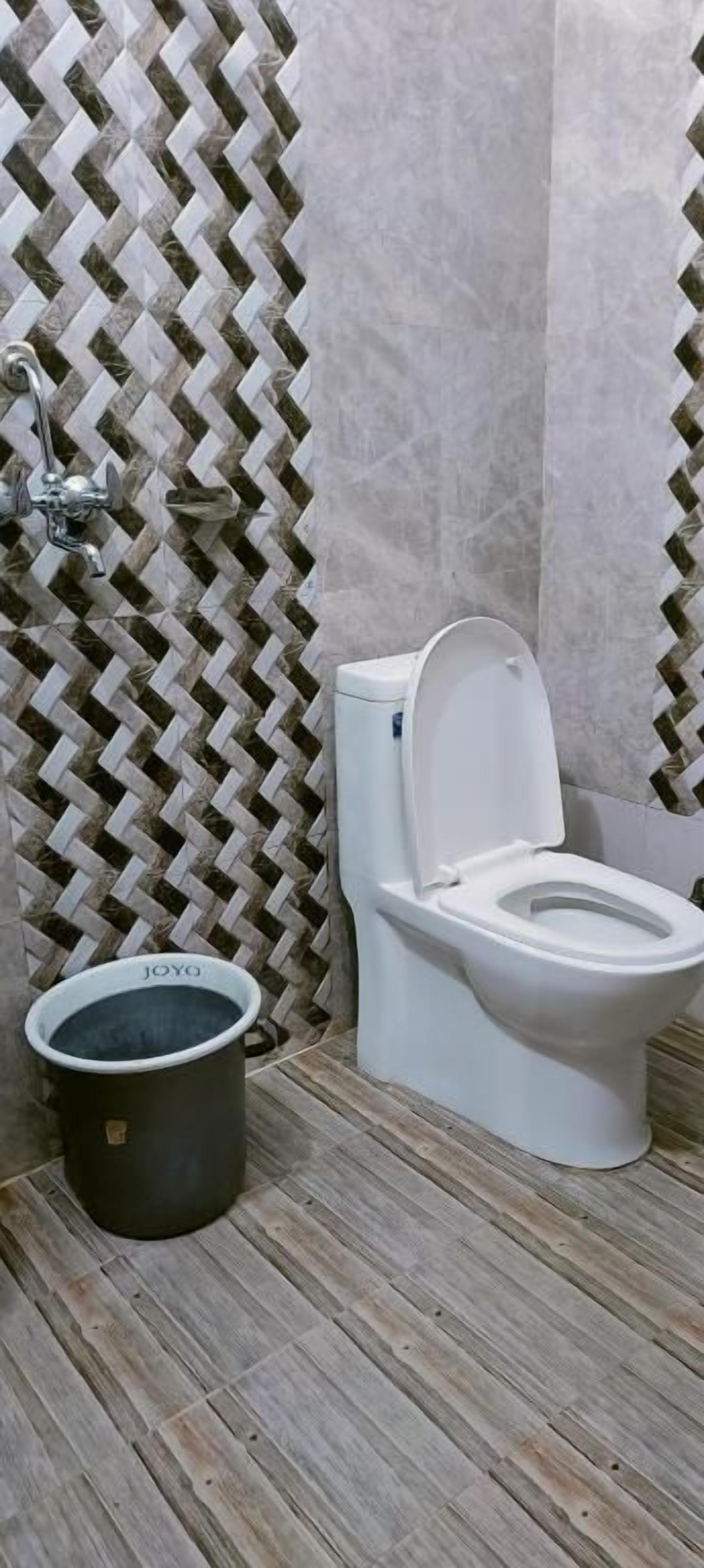 Een badkamer met een behang, een wit toilet en een zwarte emmer met de tekst 'JOYO' op de vloer.