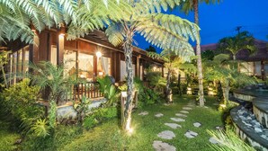 A well-lit wooden house with a garden path leading to the entrance, surrounded by various tropical plants and a small pond. - Umakelod Sebatu Villas (Sebatu)