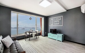 A modern living room with a large window offering a view of the sea, a sofa, a dining table with chairs, and a TV stand.