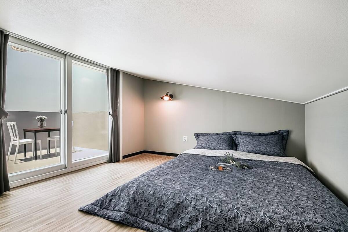 A modern hotel room with a large bed, a small table with chairs, a balcony with curtains, and a wall-mounted lamp.