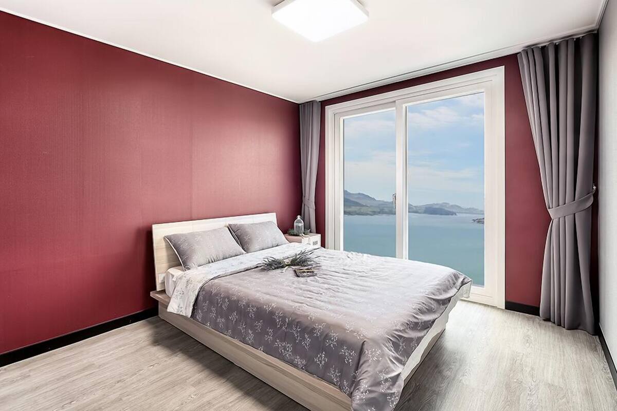 A bedroom with a large bed, a nightstand, a window with a view of the sea, and a curtain.