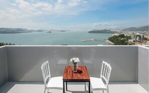 A balcony with a wooden table and two white chairs, overlooking a coastal view with buildings and a marina.