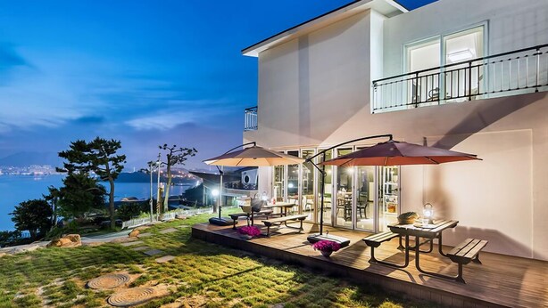 A modern house with a balcony overlooking a body of water, outdoor seating, and a well-maintained garden. - Blooming Pension (Yeosu)