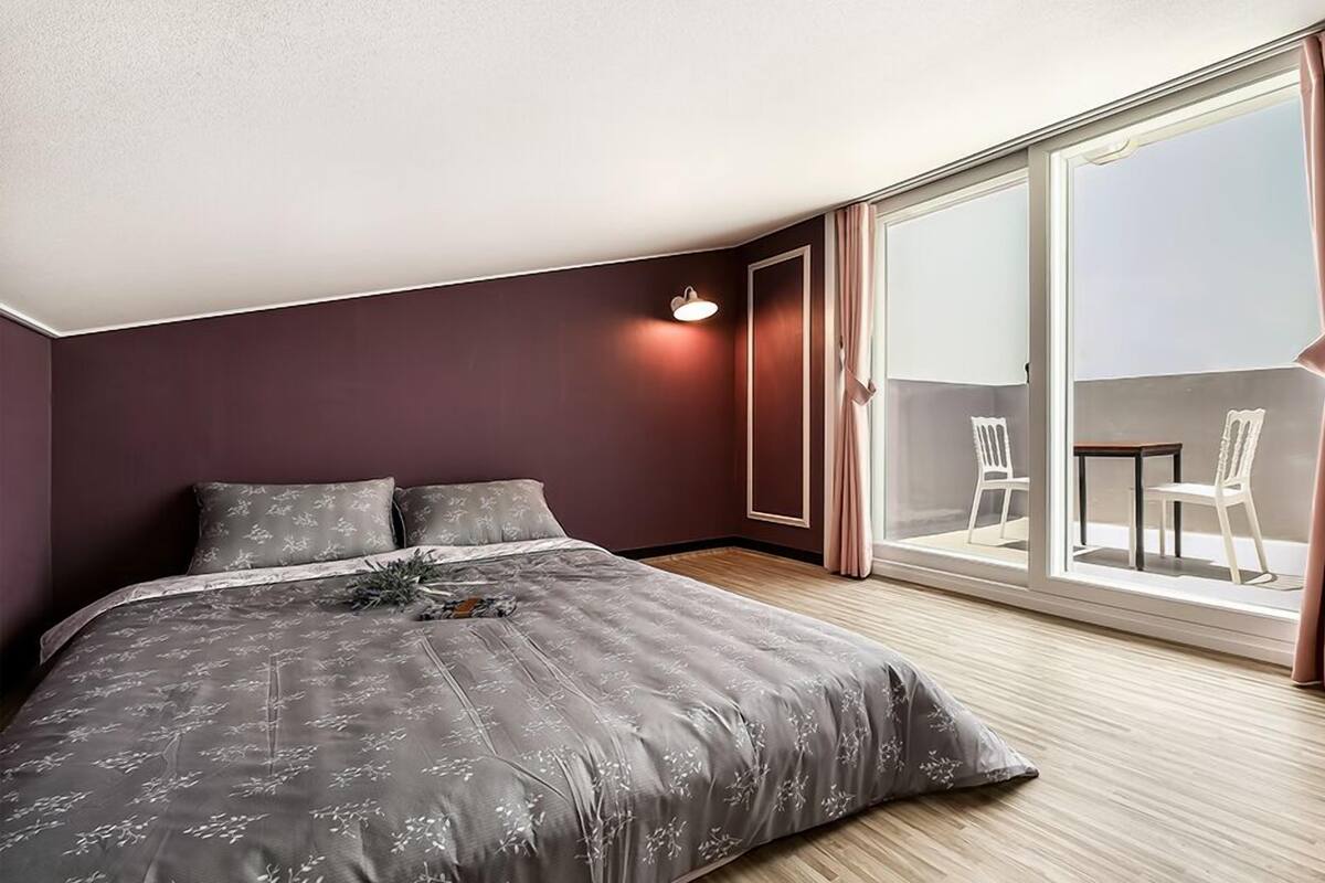 A bedroom with a large bed, a nightstand, a window with curtains, and a view of the outside balcony.