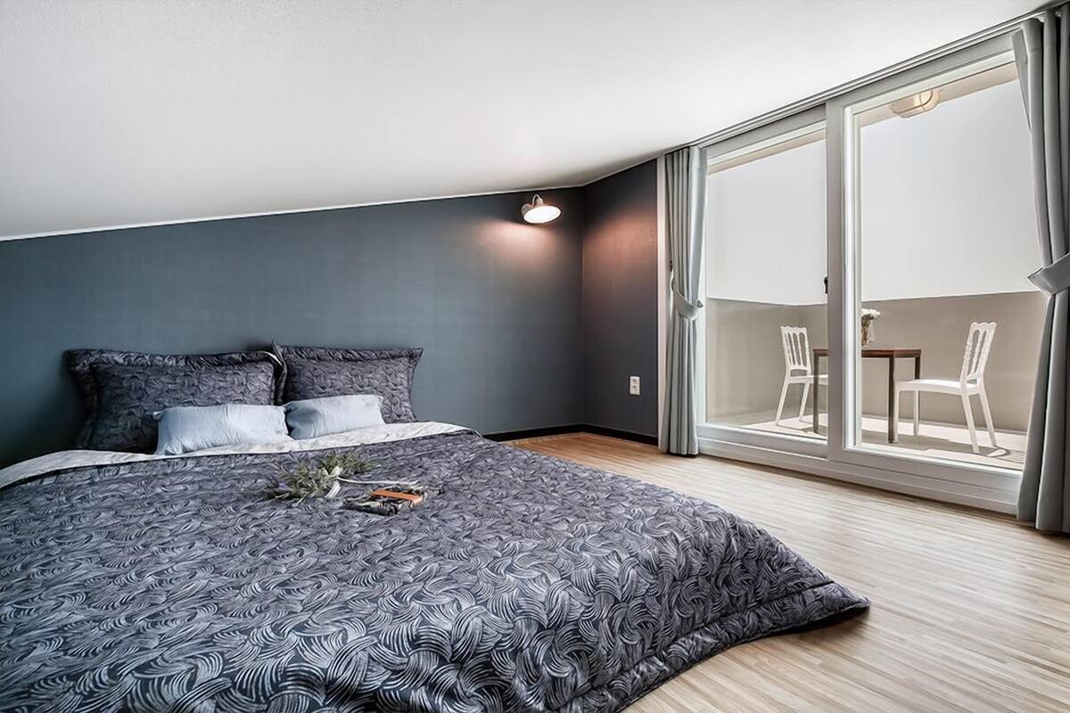 A bedroom with a large bed, a nightstand, and a window with a view of the outside.