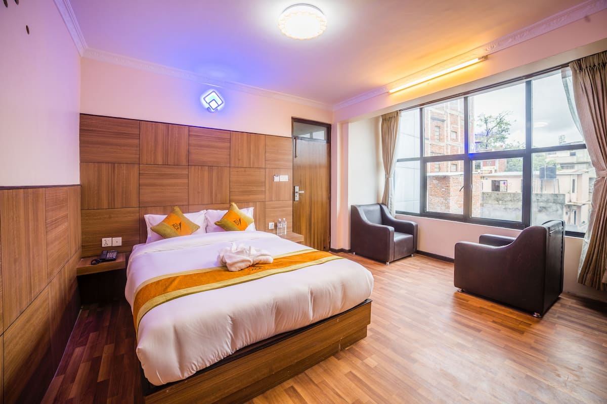 A hotel room with a large bed, a wooden headboard, a seating area with a chair, and a large window with a view of buildings.