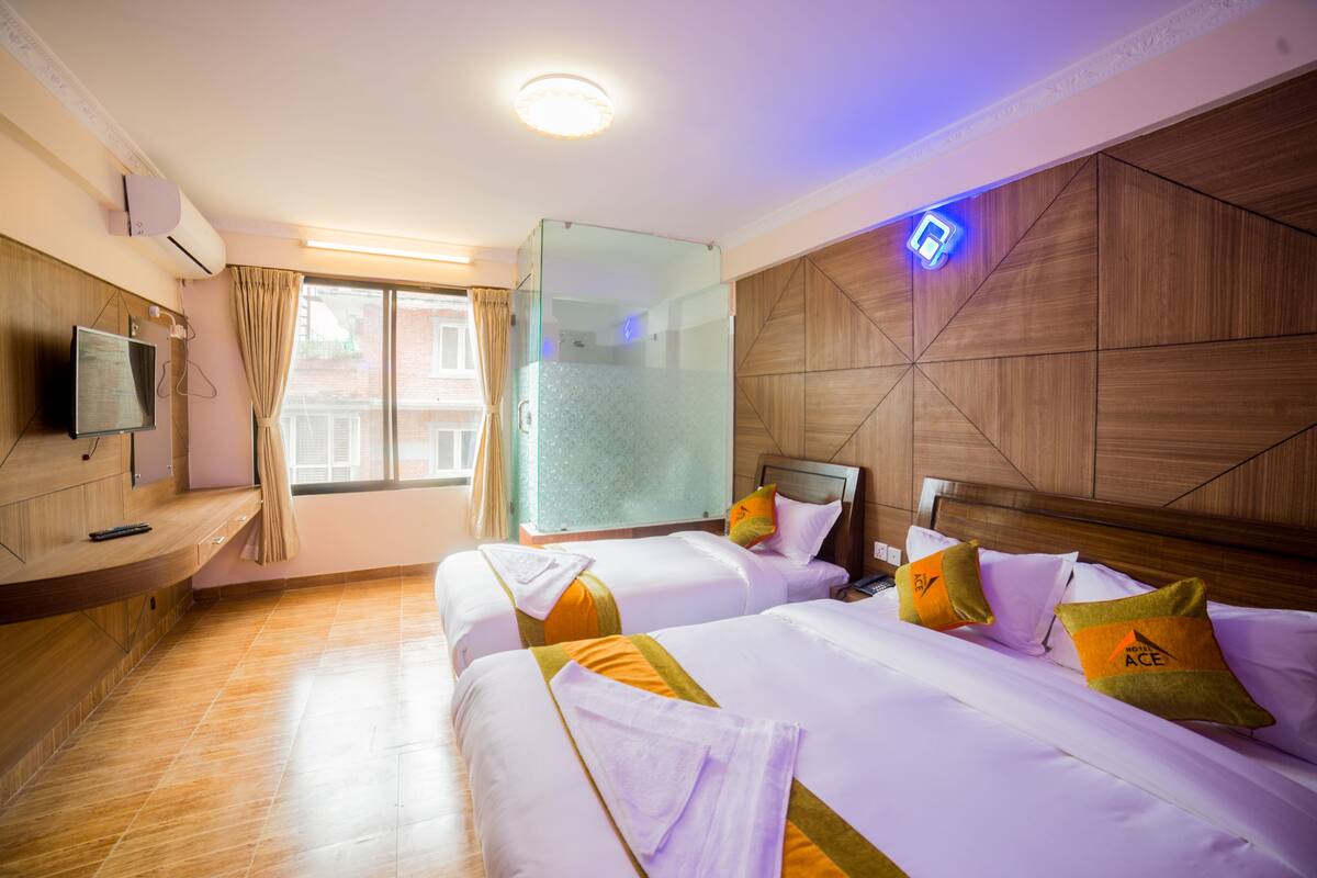 A hotel room with two beds, a TV, a glass shower enclosure, and a wooden wall panel.