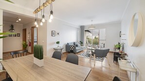 A modern living room with a dining area, featuring a wooden dining table, grey sofas, and a large window with a view.