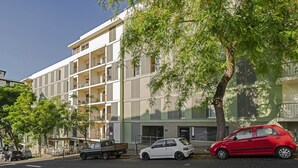 A multi-story building with balconies, a parking area, and a tree in front.