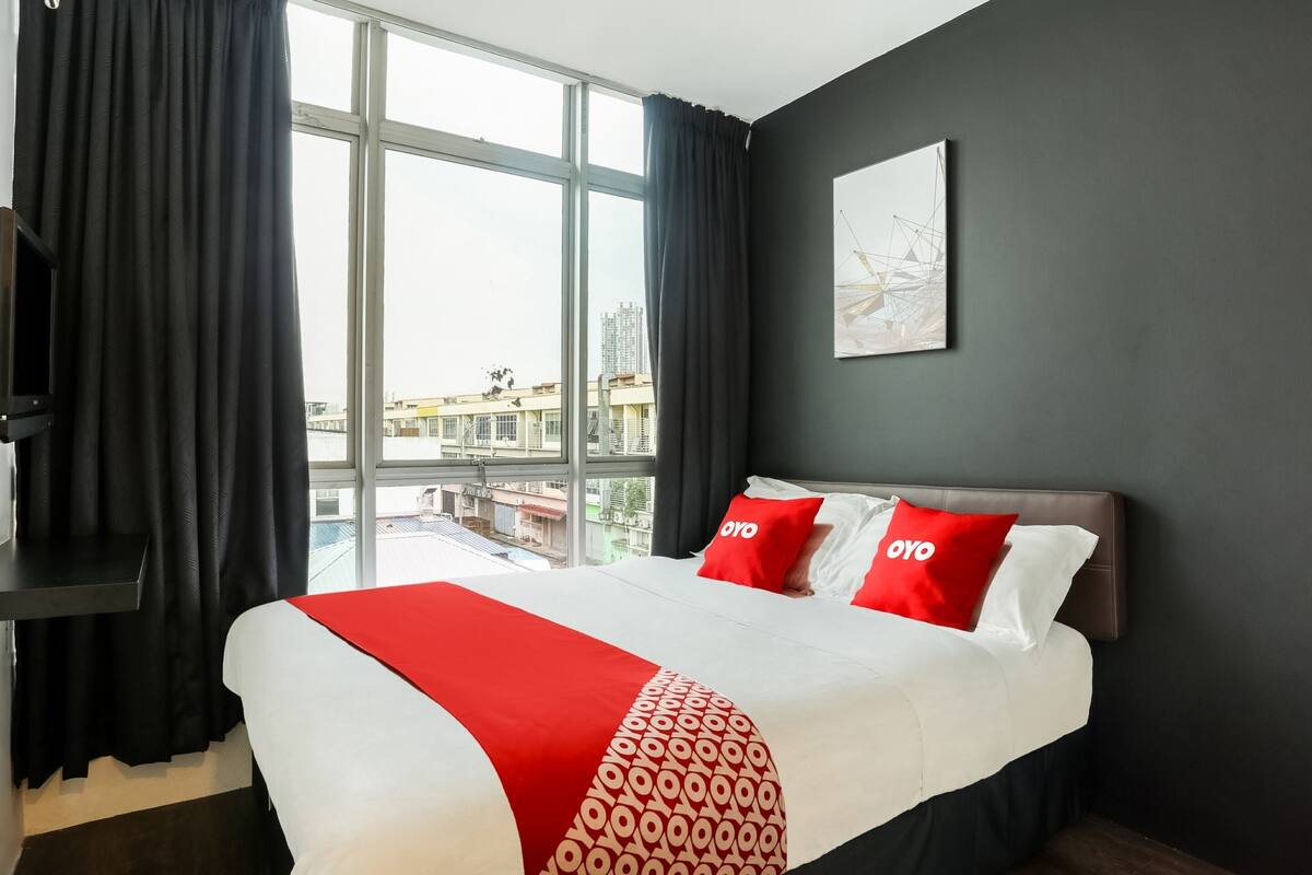 A hotel room with a large bed, black curtains, a city view, and a grey wall with a framed picture.