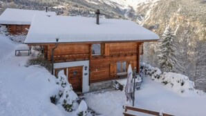 A wooden cabin with a snow-covered roof, surrounded by a snowy landscape and mountains in the background.