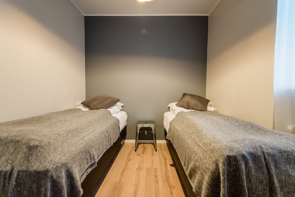 A small, well-lit room with two single beds, each with a pillow and a blanket. A small stool is placed between the beds.