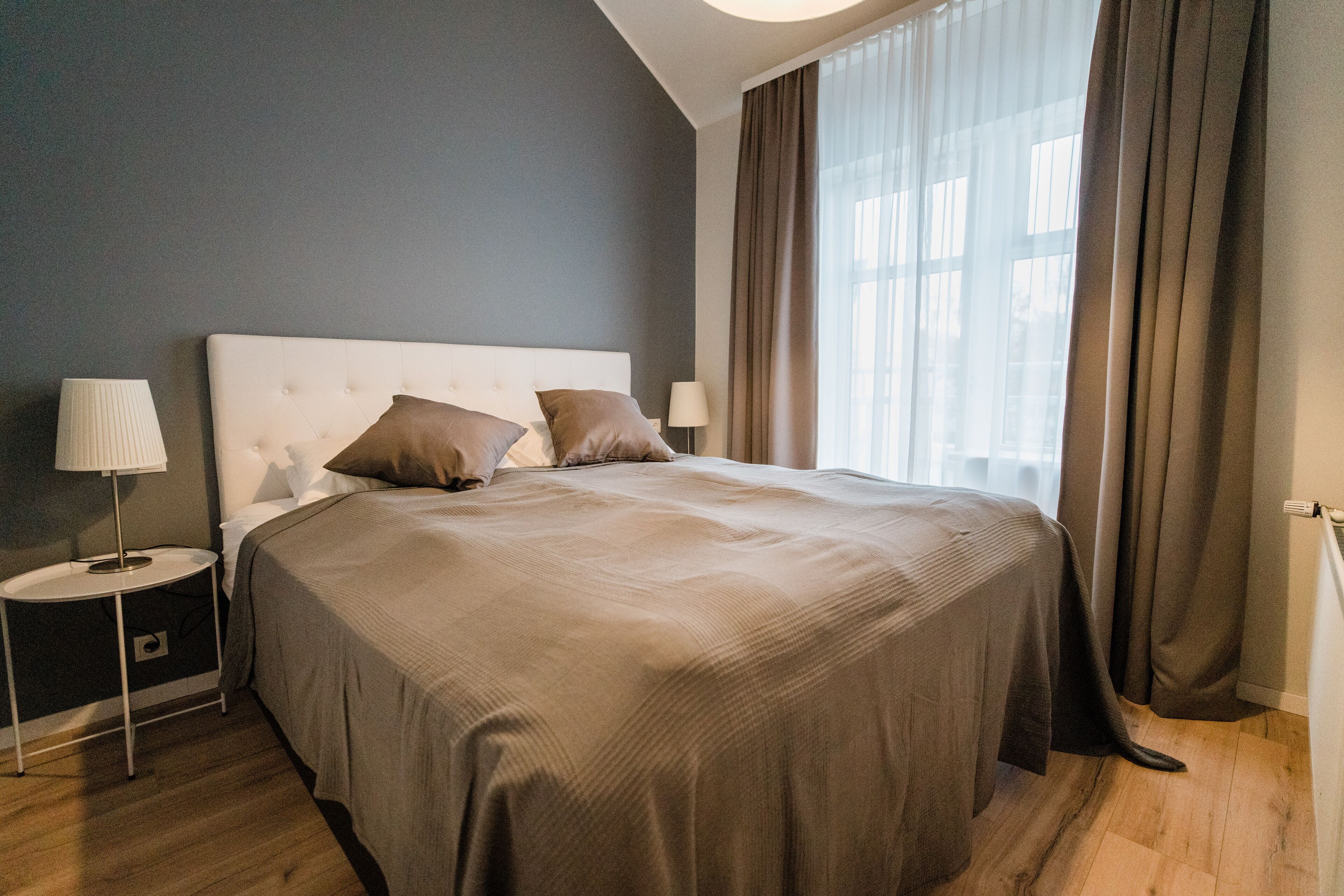 A modern hotel room with a large bed, two bedside lamps, and a view of the city through sheer curtains.